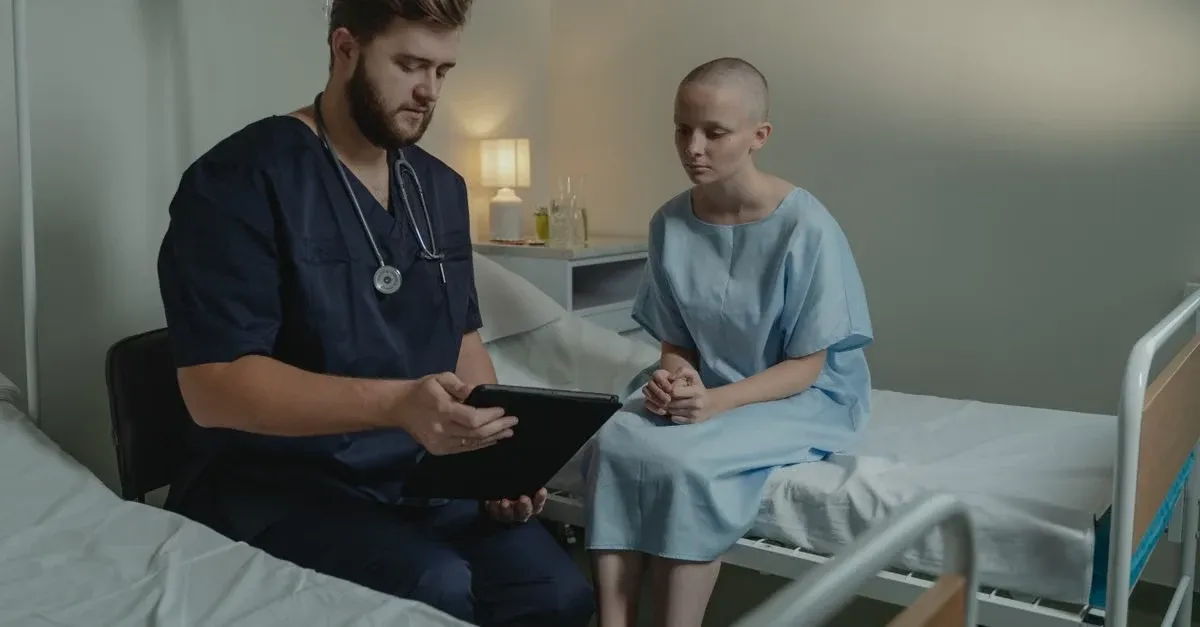 A male doctor in dark blue scrubs with a stethoscope around his neck shows a tablet to a female patient in a hospital gown sitting on a bed.