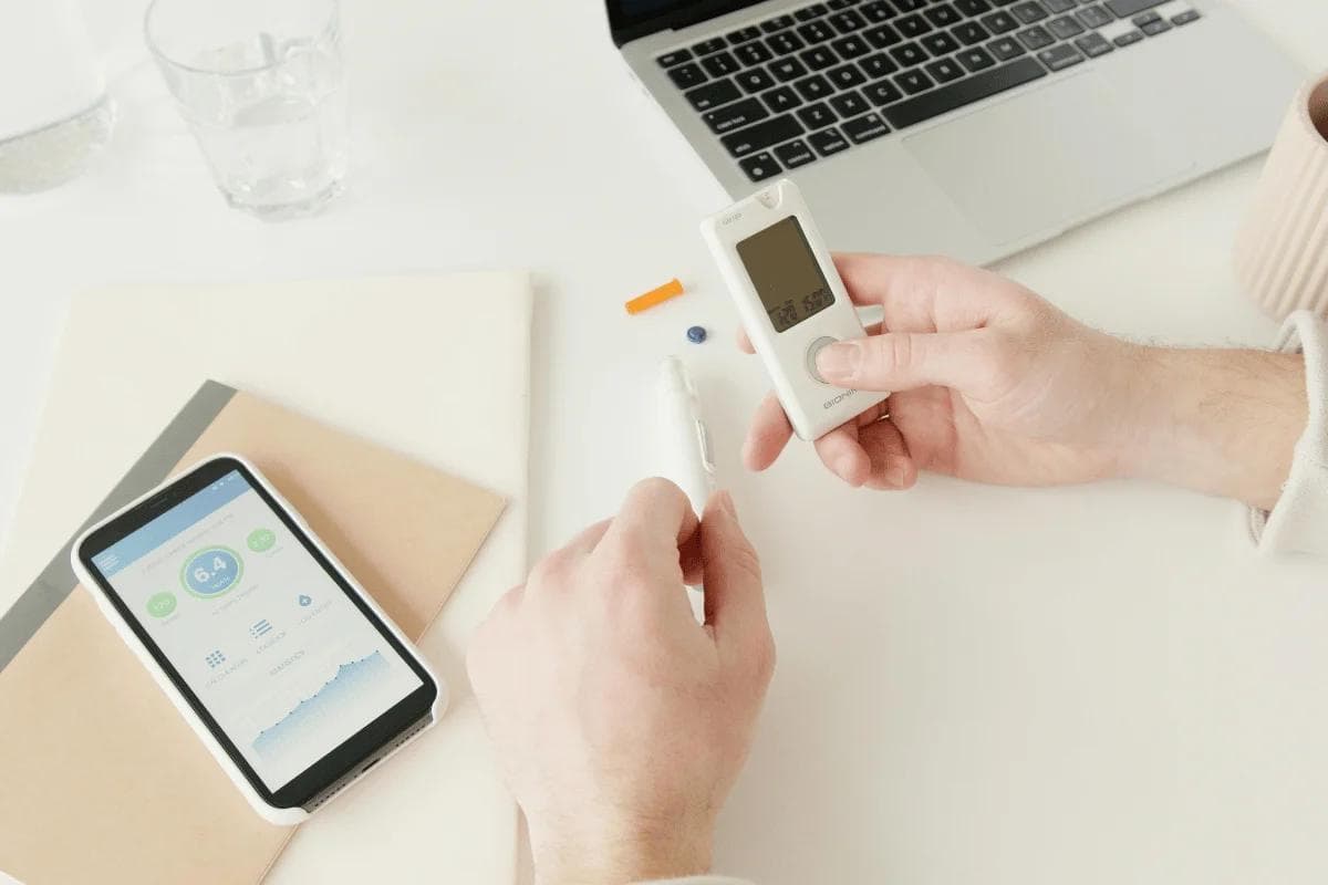 Hands holding a blood glucose meter and pen beside a smartphone displaying a health app. A laptop, notebook, and coffee cup are nearby on a white desk.