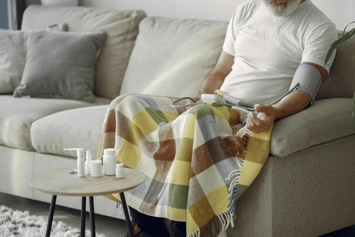 An older man checks his blood pressure while seated on a couch, covered with a colorful blanket, with medication on a nearby table.