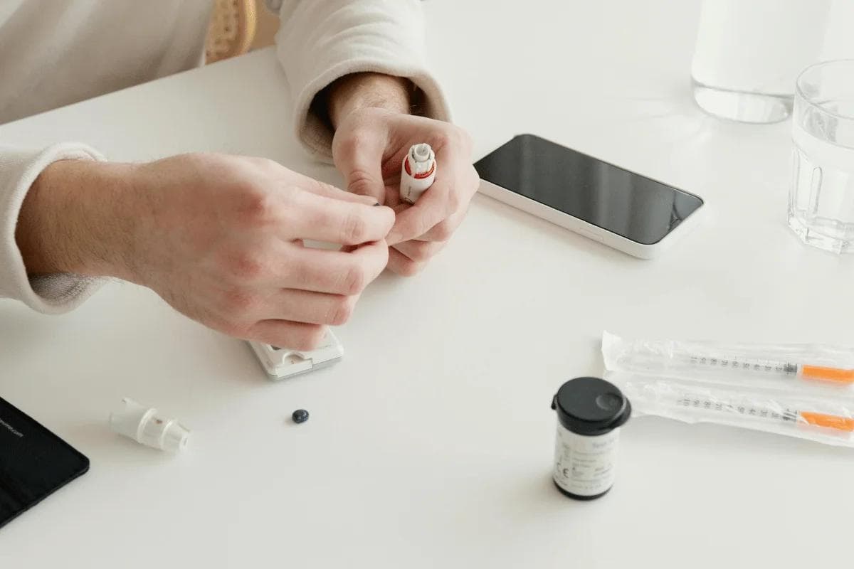 Hands assembling an insulin pen on a white table with medical supplies, a smartphone, syringes, and a glass of water nearby, conveying a medical routine.