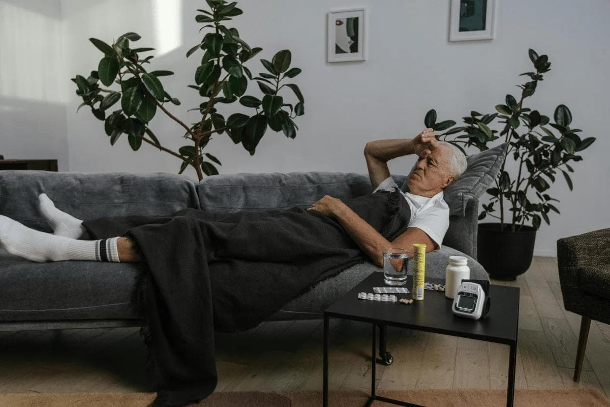 An elderly man lies on a cozy gray sofa, covered with a dark blanket, appearing unwell.