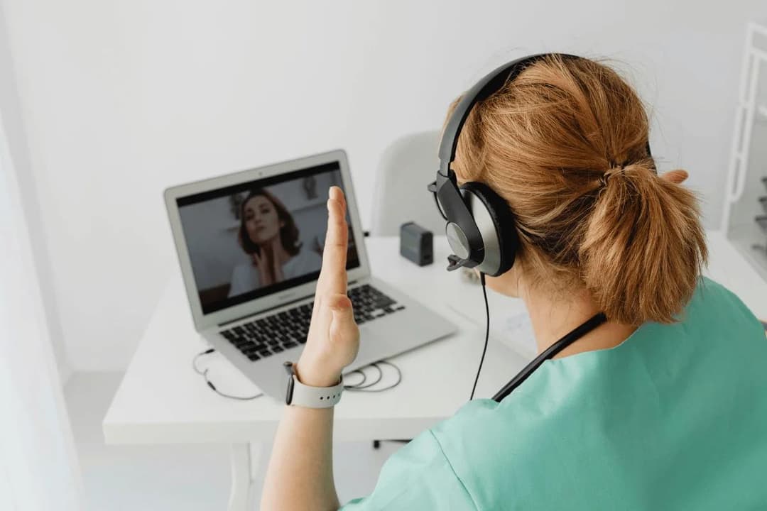 A healthcare professional in scrubs waves during a video call on a laptop. She wears headphones, engaging warmly with the person on screen.