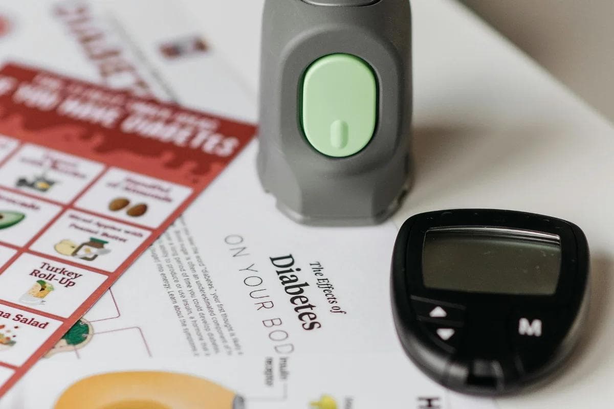 A glucose meter and a gray device with a green button sit on a brochure titled "The Effects of Diabetes on Your Body," featuring food suggestions.