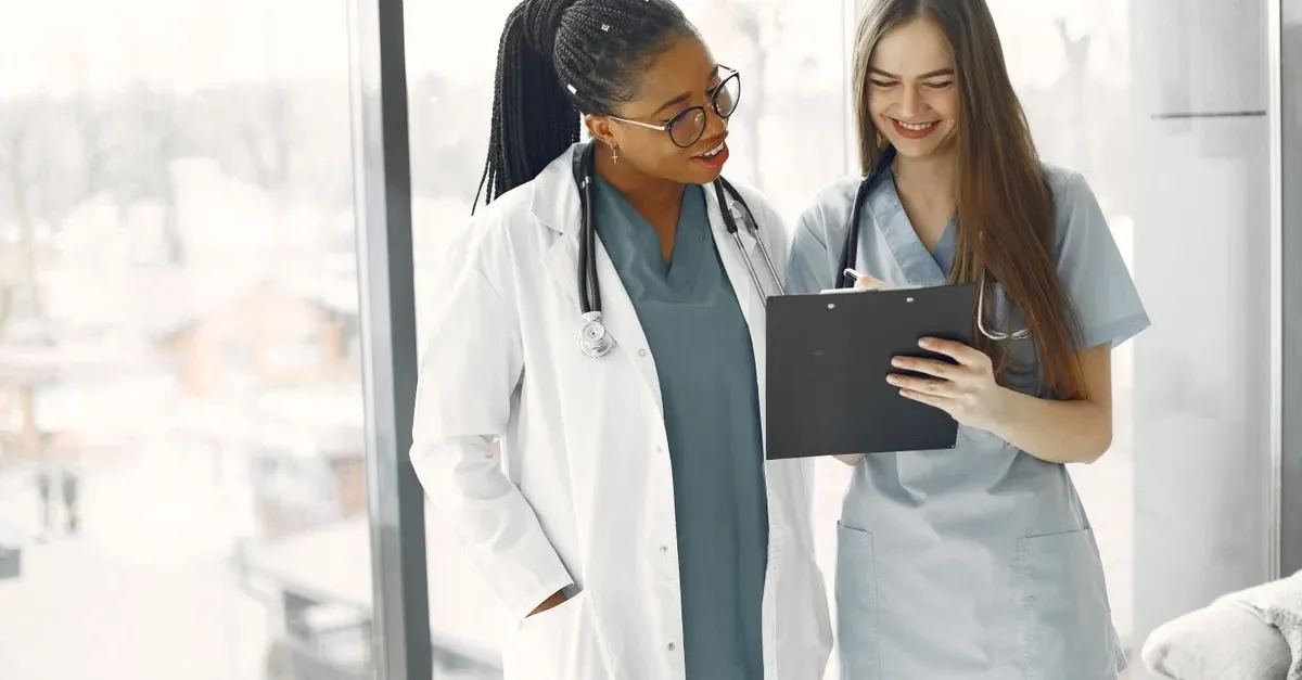 Female doctor and nurse reviewing patient records on tablet near hospital window with city skyline background.