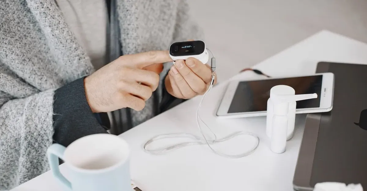 Person holding wireless health device displaying vital signs with smartphone and mobile health monitoring setup on desk.