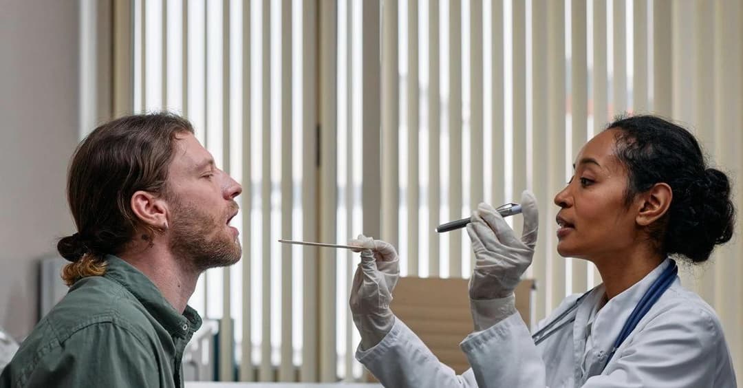 Healthcare provider conducting throat examination on patient in clinical setting with vertical blinds background.