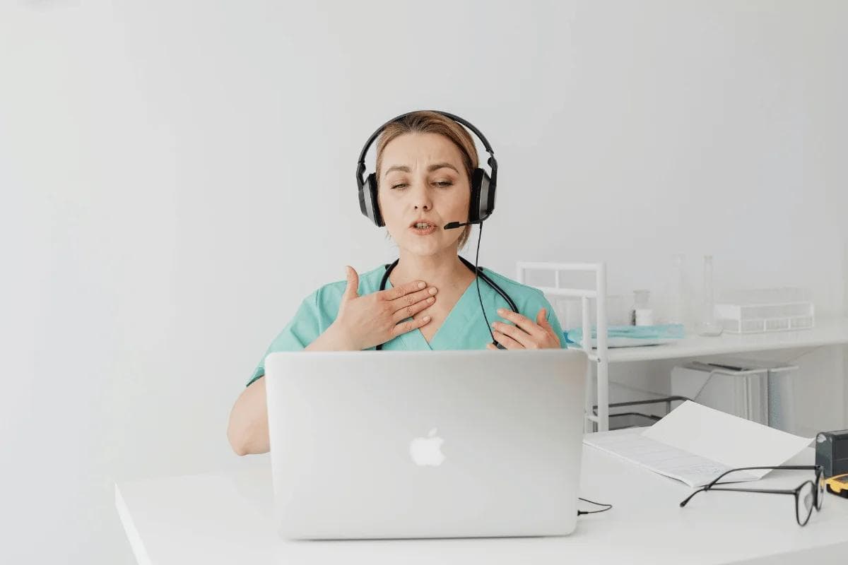 Healthcare professional in scrubs uses a headset for a telehealth video call at a desk with a laptop in a clinic office.