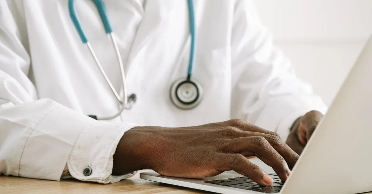 Doctor in white coat with stethoscope using laptop for rural health transformation fund applications.