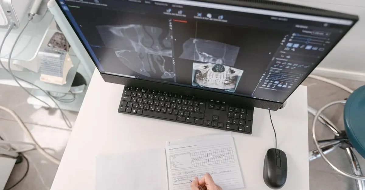 Doctor's hands reviewing patient data on form while remote patient monitoring software displays medical imaging on monitor.