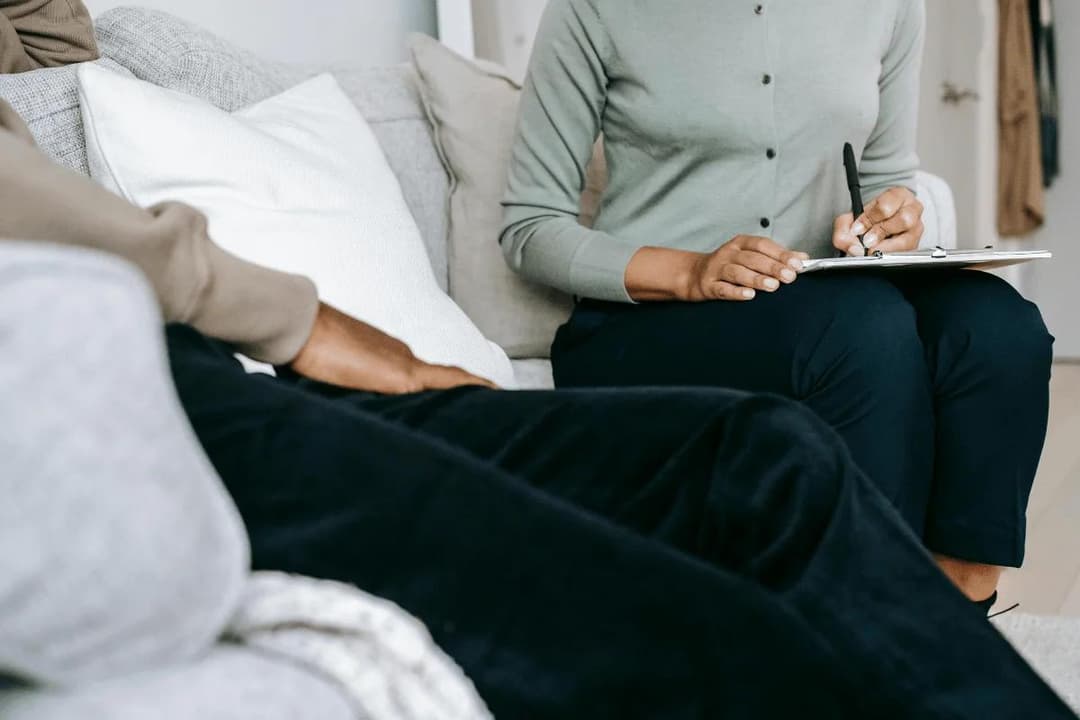 A therapist takes notes while seated on a couch, engaged in conversation with a client, who is partially visible in the foreground.